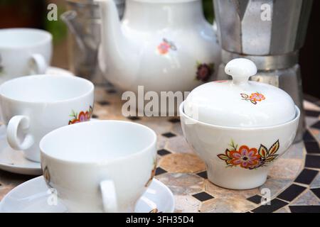 Weißes Porzellan-Teeservice mit Blumenmuster auf Mosaiktisch neben Tassen, Zuckerdose, Teekanne und Metall-Kaffeekanne in sanftem natürlichem Licht, mit Stockfoto