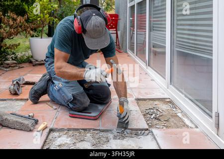Ein Arbeiter kniet auf einer Außenterrasse, während er zerbrochene Fliesen mit einem Hammer entfernt. Stockfoto