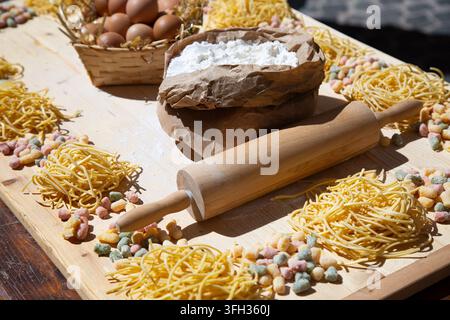 Rustikaler Backeinsatz mit Mehl in brauner Papiertüte, Holzrollenstab, Eiern im Korb, getrockneten Pastannestern und bunten Müslistücken arrangiert auf Stockfoto