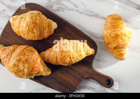 Frisch gebackene goldene Croissants, die auf einem dunklen Holzbrett über einem weißen Marmorhintergrund serviert werden Stockfoto