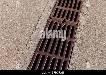 Metallgitter für Regenwasserabflusssystem, verrostete Metallgitterstäbe sind in einen Asphaltrandstein an einem sonnigen Tag eingebettet Stockfoto