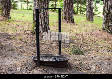 Ein einfaches schwarzes Metall-Lagerfeuer-Hängeregal steht über einem flachen Aschenbehälter auf dem Erdboden, umgeben von Kiefern. Das Lagerfeuer-Setup befindet sich in einem Stockfoto