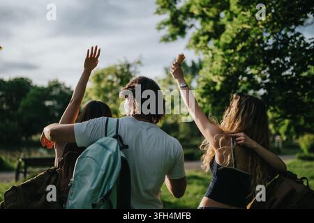Drei Studenten gehen in einem sonnigen Park mit Rucksäcken und erhobenen Armen und genießen den Sommer Stockfoto