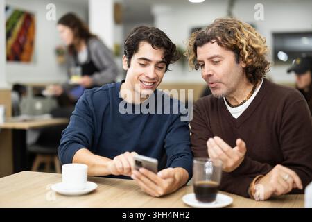 Ein glücklicher Kerl trifft sich mit Vater im Café und trinkt leckeren Kaffee und sieht sich Fotos auf dem Handy an Stockfoto