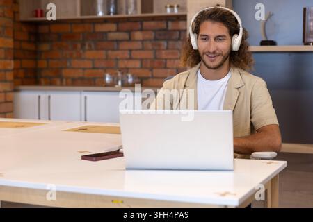 Mann sitzt an weißem Tisch in der Küche, trägt weiße Kopfhörer und benutzt Laptop und Smartphone Stockfoto
