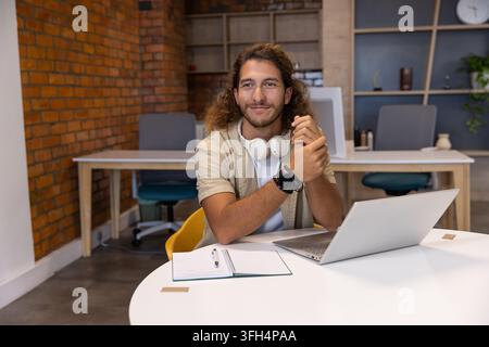 Mann sitzt an einem runden weißen Tisch in einem Co-Working-Studio und verwendet einen silbernen Laptop, ein Notizbuch und Kopfhörer Stockfoto