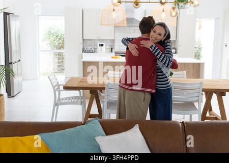 Paar umarmt sich, Frau lächelt in gestreiftem Oberteil, Mann trägt rote Jacke an Holz-Esstisch Stockfoto