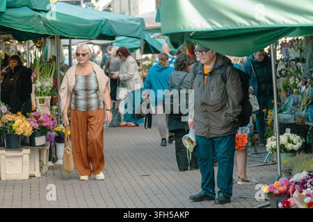 RIGA, LETTLAND - 7. SEPTEMBER 2026: Einkäufer gehen zwischen bunten Blumenständen auf einem belebten Markt im Freien umher Stockfoto