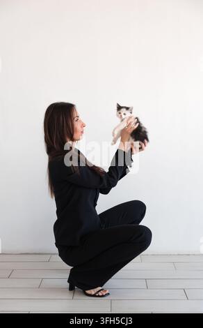 Frau in schwarzem formellen Hosenanzug sitzt an weißer Wand und hält ein kleines süßes Kätzchen. Kaukasisches brünettes Model mit langen Haaren. Geschäftsporträt Stockfoto
