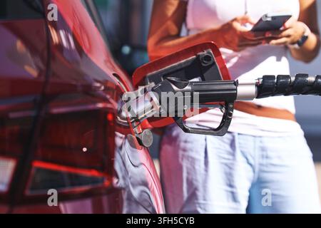 Frau steht neben rotem Auto und tankt an einer Tankstelle, überprüft das Telefon während steigender Kraftstoffpreise und Inflation Stockfoto