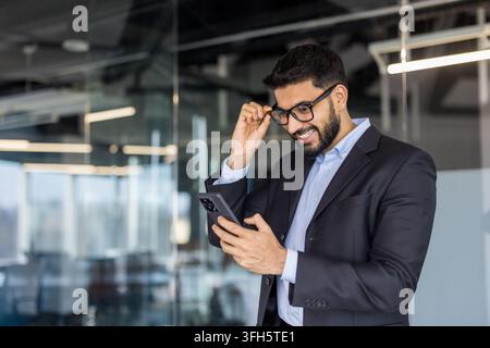 Digitaler Wandel und Geschäftskommunikationskonzept, lächelnder mittelöstlicher Geschäftsmann verwendet Smartphone im modernen Büro. Professioneller Manager überprüft Mobilgerät und passt Brille an. Stockfoto
