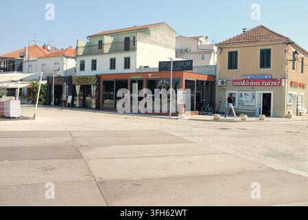 Pirovac, Kroatien, 16. August 2026: Sonnige, helle Küstengegend mit niedrigen Ziegeldächern, Ladenfronten und kleinen Restaurantfassaden entlang gepflasterter Wege Stockfoto