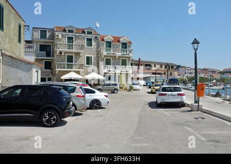 Pirovac, Kroatien August 16, 2026 Parkte Autos säumen die schmale Uferstraße neben einem Steinhaus mit grünen Fensterläden, Balkonen und einem roten Ziegeldach Stockfoto