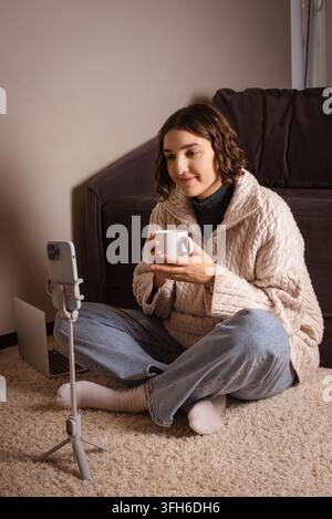 Junge weibliche Content-Erstellerin hält eine Kaffeetasse und nimmt einen Videoblog auf einem Smartphone auf, das auf einem Stativ montiert ist Stockfoto