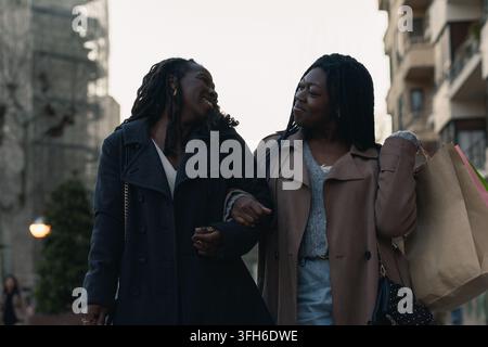 Zwei fröhliche afrikanische Frauen schlendern Arm in Arm und schauen sich mit Komplizenschaft durch die Straßen der Stadt an, genießen einen gemeinsamen Shoppingtag Stockfoto