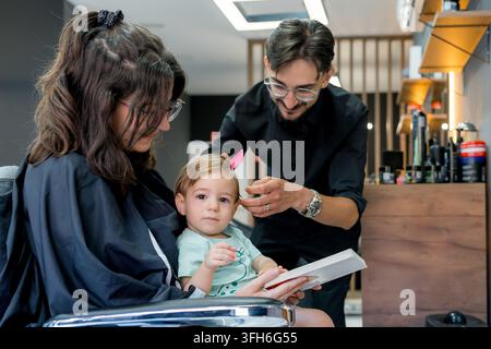 Professioneller Friseur gibt einem Kleinkind einen sorgfältigen Haarschnitt in einem modernen Friseursalon, während das Kind auf dem Schoß der Mutter sitzt, und schafft so eine ruhige Familiengrooming-Umgebung für die Mutter Stockfoto