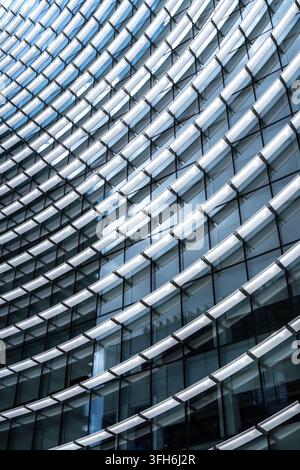 Nahaufnahme eines zeitgenössischen Bürogebäudes mit einer geschwungenen geometrischen Fassade, wiederholten Glasfenstern und silbernen Metall-Lamellen, die ein Abstraktes schaffen Stockfoto