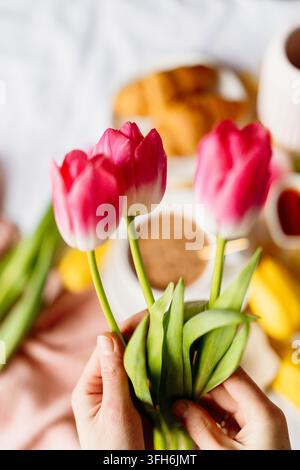 Nahaufnahme von Händen, die einen frischen Strauß rosa Tulpen über einem gemütlichen Frühstückstisch mit Brot, Kaffee, Obst und weichem natürlichem Licht in einem hellen Ho halten Stockfoto