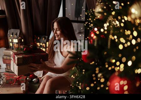 Eine junge Frau hält verpackte Weihnachtsgeschenke in der Nähe eines geschmückten Baumes an einem gemütlichen Feiertagswinterabend. Stockfoto