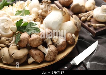 Viele verschiedene Pilze, Petersilie und Messer auf Holztablett, Nahaufnahme Stockfoto
