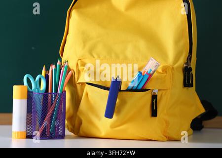 Rucksack mit Schreibwaren auf weißem Schreibtisch vor Kreidetafel, Nahaufnahme Stockfoto