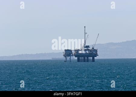 Große Offshore-Ölplattform ragt hoch in ruhiger blauer See. Ferne Hügel verschwinden im dunstigen Horizont unter klarem Himmel. Industrielle Struktur wirkt isoliert, dürr Stockfoto