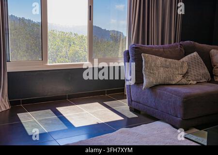 Sonnenbeleuchtetes modernes Wohnzimmerinterieur mit großem Fenster und dunklen Vorhängen Stockfoto
