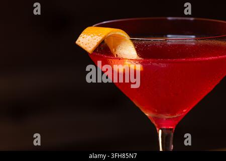 Rosa Cocktail im Coupe-Glas. Eine Orangenschneute schmückt den Rand. Dunkler Holztisch und dunkler Hintergrund schaffen eine rustikale Baratmosphäre. Stockfoto