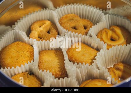 Hochwinklige Nahaufnahme verschiedener Formen von goldenen Butterkeksen, die in einzelnen weißen Papierbechern in einer Metallblechdose angeordnet sind. Stockfoto