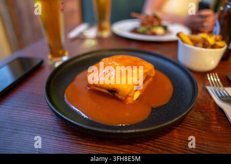 Nahaufnahme eines veganen Francesinha-Sandwiches, bedeckt mit geschmolzenem Käse und Sauce, serviert mit einer Beilage Pommes und einem Glas Bier auf einem Holztisch in einem Li Stockfoto
