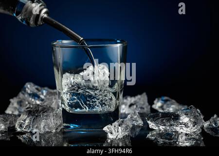 Kristallklarer Wodka oder Gin wird über Eis in ein Glas gegossen. Wodka und Eisstücke auf schwarzem reflektierendem Hintergrund. Kopierbereich. Stockfoto