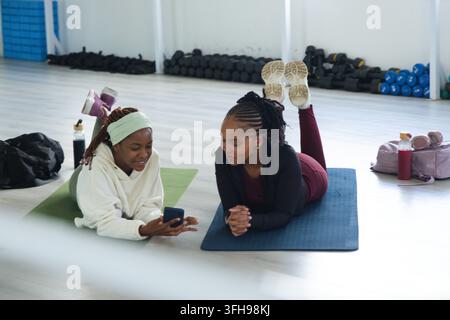 Zwei Frauen liegen auf Yogamatten im Fitnessstudio, tragen Sportkleidung und zeigen ein Smartphone mit Wasserflaschen in der Nähe Stockfoto