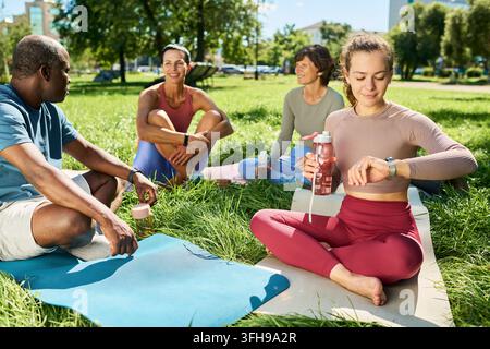 Frau, die eine Smartwatch überprüft, sitzt mit einer vielfältigen Gruppe auf Matten im Park Stockfoto