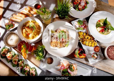 Eine Top-Down-Ansicht einer Vielzahl von thailändischen Hauptgerichten auf einem Esstisch, darunter Pad Thai, Panang-Curry, Hühnchensatay, grünes Curry und Garnelen-Miang Kham. Stockfoto