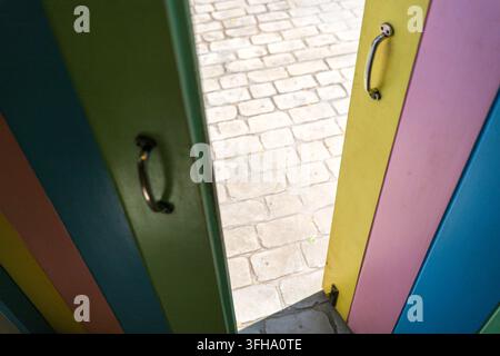 Bunte Holzinnentüren öffnen sich zu einem sonnenbeschienenen Ziegelweg und schaffen einen einladenden Eingang mit kräftig gestreiften Farben und geschwungenen Metallgriffen im Spielbereich Stockfoto