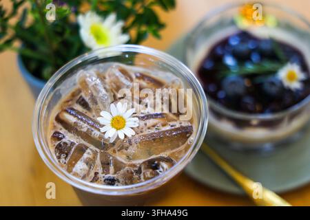 Ein Eiskaffee mit Eiswürfel und Gänseblümchengarnitur neben Blaubeerdessert und Blume auf Holztablett, die ein erfrischendes Café-Stillleben mit natürlichem l Stockfoto