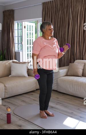 Senior Afroamerikanerin verwendet lila Hanteln auf einer Yogamatte mit Flasche im Wohnzimmer Stockfoto