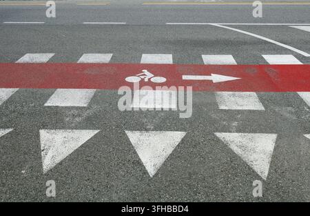 Rotes Fahrradweg-Schild entlang eines Zebrastreifens, mit einem gemalten Vorfahrtszeichen vorne. Urbane Straße im Hintergrund. Stockfoto