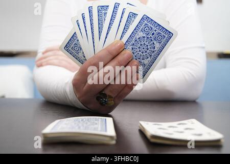 Nahaufnahme einer Person, die ein Fächer blaubackiger Spielkarten bei einem Kartenspiel an einem Tisch hält, mit zusätzlichen Karten und einem Kartenspiel im Vordergrund. Stockfoto