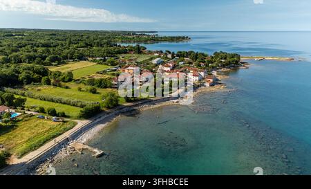 Blick auf ein friedliches Küstendorf mit roten Dächern, felsiger Küstenlinie und klarem türkisfarbenem Wasser, umgeben von grünen Feldern und Wald, Umag, Kroatien Stockfoto