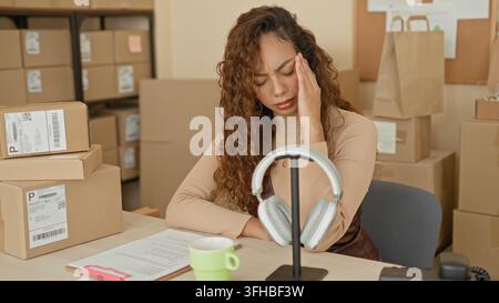 Junge hispanische Frau mit Hand zum Tempel, die Paketpapiere neben gestapelten Versandkartons im Gebäude liest; Stressbestandsmanagement. Stockfoto