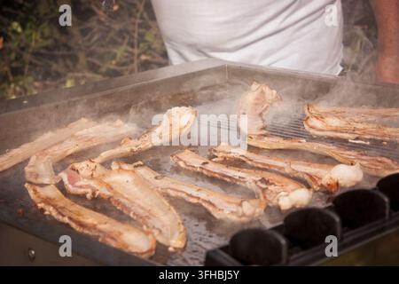 Speckscheiben brutzeln auf einem Flächengrill im Freien, verströmen rauchigen Duft und werden bei einem lockeren Barbecue oder einem Grillfest im Garten goldbraun knusprig Stockfoto