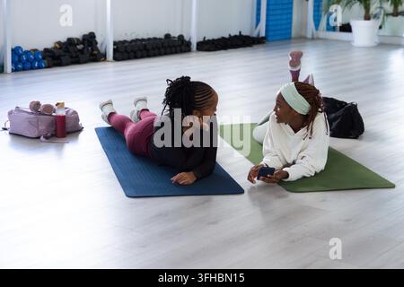Zwei erwachsene afroamerikanische Frauen ruhen auf Matten im Studio, halten ein Smartphone neben einer rosa Sporttasche Stockfoto