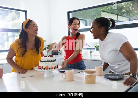 Verschiedene Freundinnen lachen, schneiden auf einer weißen Säule auf der Kücheninsel einen mehrschichtigen Kuchen ab, mit Tassen Stockfoto
