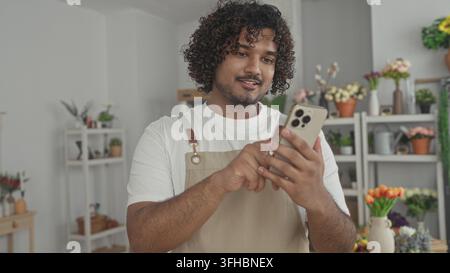 Mann Florist klopft Smartphone mit Händen in einem Blumenladen Gebäude, trägt eine Schürze und lächelt auf den Bildschirm; kleine Unternehmen kümmern sich gut. Stockfoto