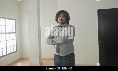 Mann mit überkreuzten Armen, gepunktetem Hemd und Krawatte, stehend in einem leeren Bauraum an einem großen Fenster und Betonsäule; Vertrauen. Stockfoto