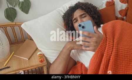 Mann im Bett mit Smartphone, Hand am Hals und Griff nach der Pille Flasche auf dem Nachttisch, bedeckt mit orangefarbener Decke, Studio; krank. Stockfoto