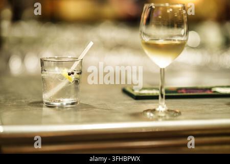 Gin- und Weißweingläser auf einer polierten Theke in einer dämmrigen Lounge, Gin Tonic mit Zitrone und Pfefferkörnern neben einem gekühlten Glas Weißwein, e Stockfoto