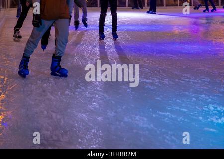 Nahaufnahme von Beinen mit blauen Eisschuhen, die auf einer strukturierten Eisfläche gleiten, beleuchtet von dynamischem violettem und orangefarbenem Licht, symbolisch für saisonale Freude und Aktivität Stockfoto