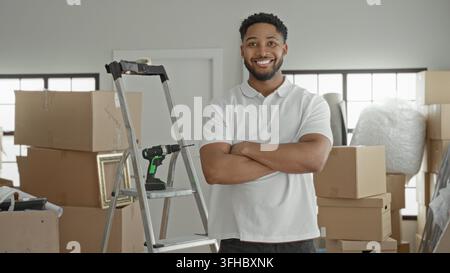 Mann mit Kartons und Akku-Bohrer, Arme verschränkt neben einer Leiter im Gebäude; Selbstvertrauen am Umzugstag. Stockfoto